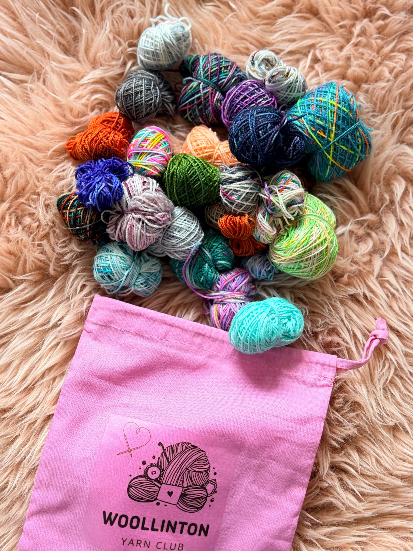 A pink Woollinton Yarn Club project bag overflowing with a colourful variety of hand-dyed fingering weight mini skein yarn balls on a fluffy neutral background.